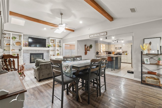 dining area featuring visible vents, a brick fireplace, ceiling fan, vaulted ceiling with beams, and light wood finished floors