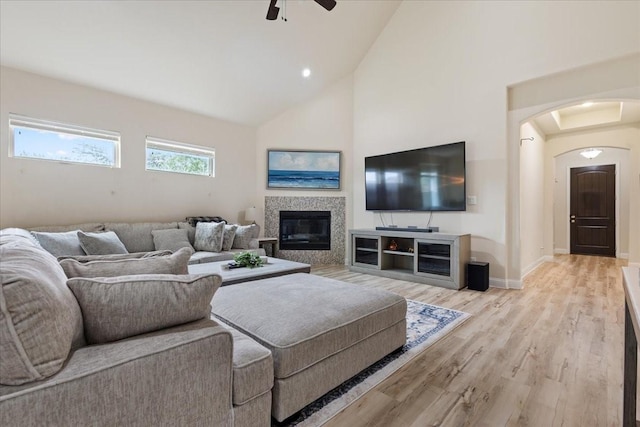 living area featuring light wood-style floors, ceiling fan, high vaulted ceiling, a tiled fireplace, and arched walkways