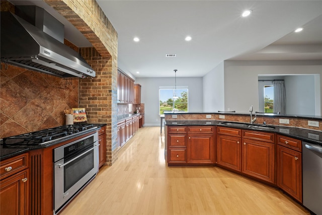 kitchen with appliances with stainless steel finishes, tasteful backsplash, a sink, light wood finished floors, and wall chimney exhaust hood