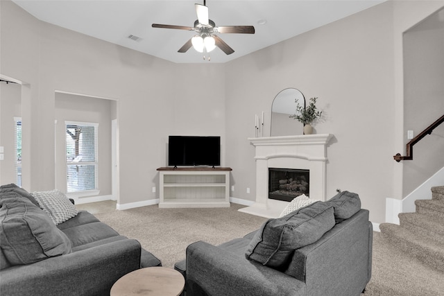 living room featuring a fireplace with raised hearth, ceiling fan, visible vents, stairs, and carpet floors