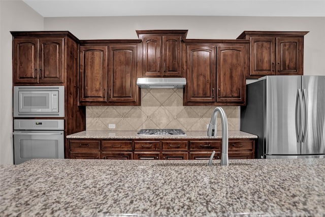 kitchen with stainless steel appliances, under cabinet range hood, backsplash, light stone countertops, and dark brown cabinets