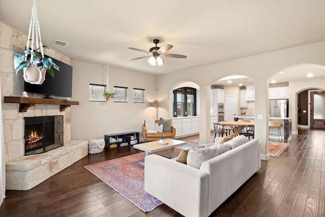 living room featuring ceiling fan, a stone fireplace, visible vents, arched walkways, and dark wood-type flooring