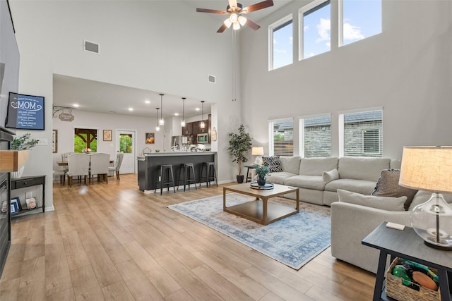 living room featuring a healthy amount of sunlight, ceiling fan, visible vents, a high ceiling, and light wood-style flooring