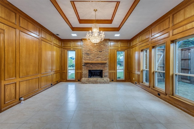 unfurnished living room featuring crown molding, a notable chandelier, a raised ceiling, a decorative wall, and a brick fireplace