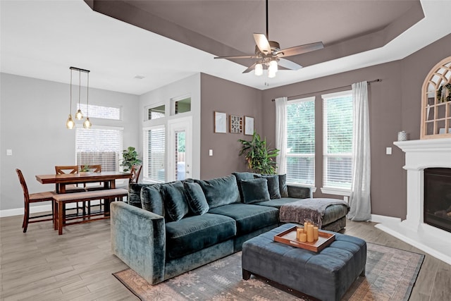 living room featuring light wood-style floors, baseboards, a ceiling fan, a glass covered fireplace, and a tray ceiling