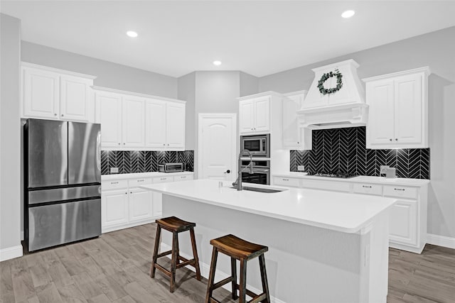 kitchen with appliances with stainless steel finishes, a sink, premium range hood, white cabinets, and a kitchen island with sink