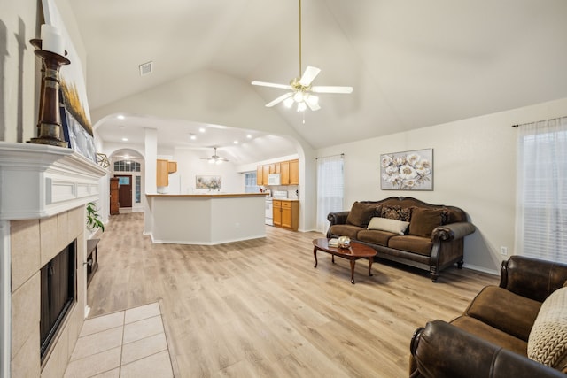 living area featuring ceiling fan, arched walkways, a tile fireplace, high vaulted ceiling, and light wood finished floors