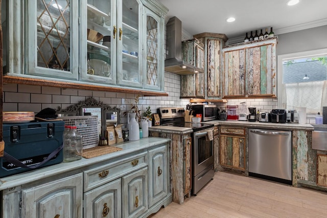 kitchen featuring decorative backsplash, glass insert cabinets, light wood-style floors, wall chimney exhaust hood, and stainless steel appliances