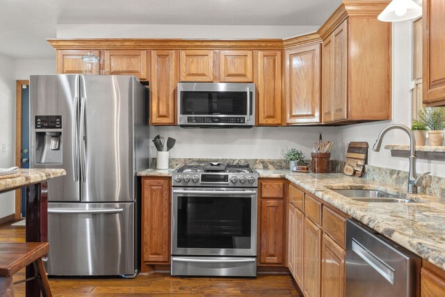 kitchen featuring brown cabinetry, dark wood-style flooring, light stone countertops, a sink, and appliances with stainless steel finishes