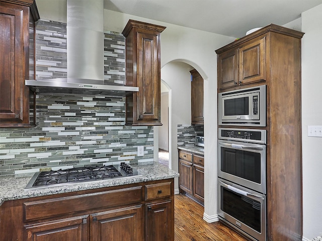 kitchen featuring decorative backsplash, dark wood-style flooring, wall chimney range hood, arched walkways, and appliances with stainless steel finishes
