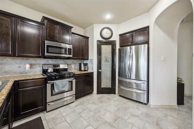 kitchen featuring dark brown cabinetry, baseboards, tasteful backsplash, light stone counters, and appliances with stainless steel finishes