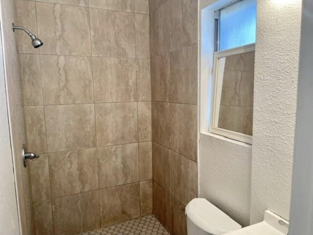 full bath featuring toilet, a textured wall, and tiled shower