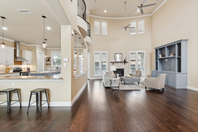 living room with dark wood-style flooring, ceiling fan, a stone fireplace, visible vents, and ornamental molding