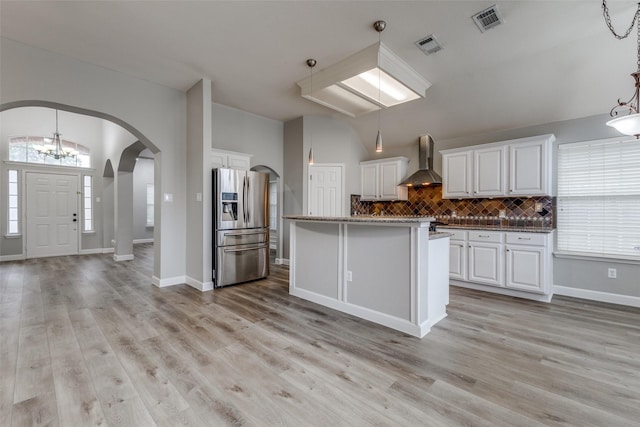 kitchen with stainless steel refrigerator with ice dispenser, arched walkways, tasteful backsplash, white cabinetry, and wall chimney exhaust hood