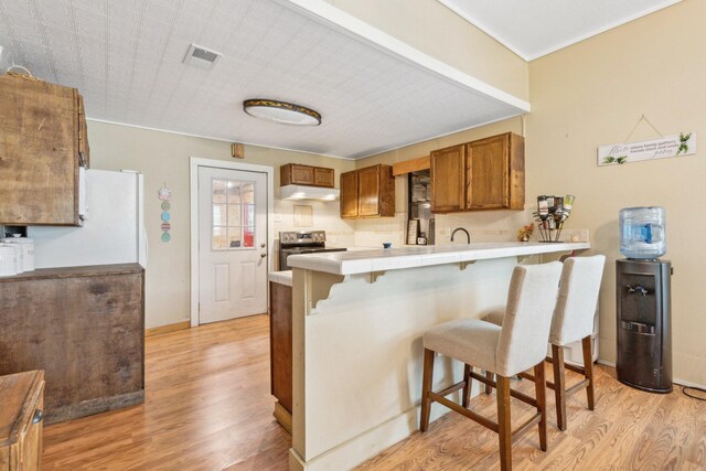 kitchen with brown cabinetry, a peninsula, light wood-type flooring, a kitchen bar, and under cabinet range hood