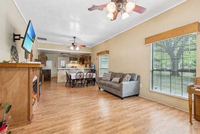 living room with crown molding, light wood-style floors, a glass covered fireplace, a ceiling fan, and baseboards