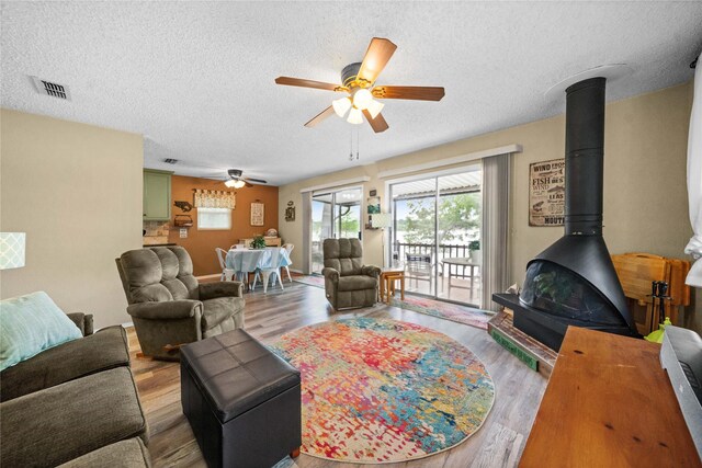 living room featuring visible vents, a wood stove, wood finished floors, a textured ceiling, and a ceiling fan