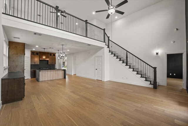 unfurnished living room with visible vents, a towering ceiling, light wood-type flooring, ceiling fan with notable chandelier, and stairs