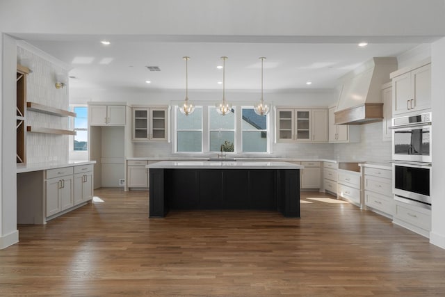 kitchen featuring a kitchen island, backsplash, stainless steel double oven, custom exhaust hood, and glass insert cabinets