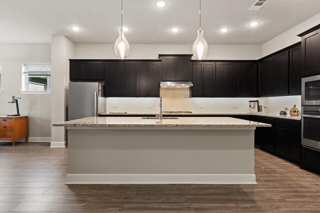 kitchen featuring appliances with stainless steel finishes, light stone countertops, a sink, dark wood-style floors, and an island with sink