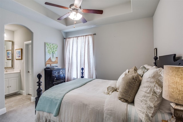 bedroom with baseboards, light colored carpet, a raised ceiling, connected bathroom, and arched walkways