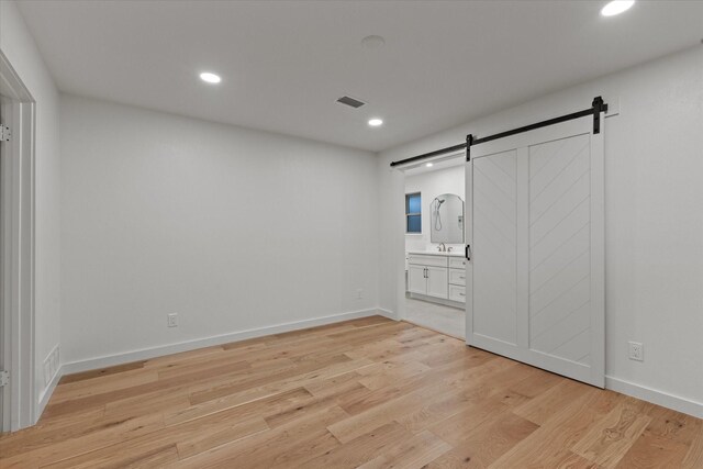 unfurnished bedroom featuring a barn door, light wood finished floors, recessed lighting, ensuite bathroom, and baseboards