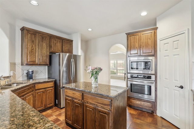 kitchen with appliances with stainless steel finishes, a sink, arched walkways, dark stone countertops, and tasteful backsplash