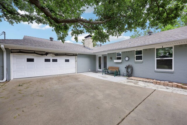 rear view of house featuring roof with shingles, an attached garage, brick siding, a chimney, and concrete driveway