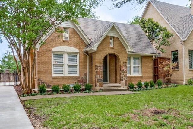 tudor home featuring fence, a front yard, roof with shingles, and brick siding