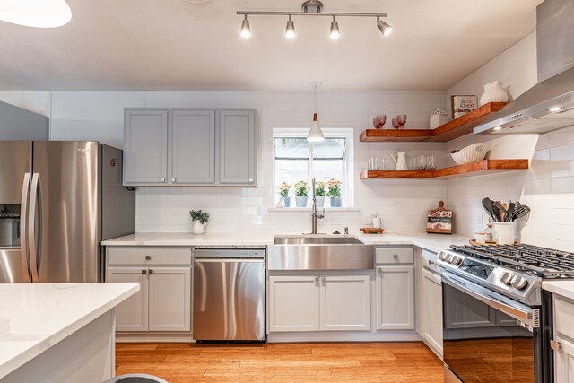 kitchen featuring wall chimney range hood, appliances with stainless steel finishes, a sink, light wood finished floors, and backsplash