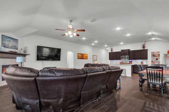 living area with a ceiling fan, visible vents, a tiled fireplace, vaulted ceiling, and dark wood finished floors