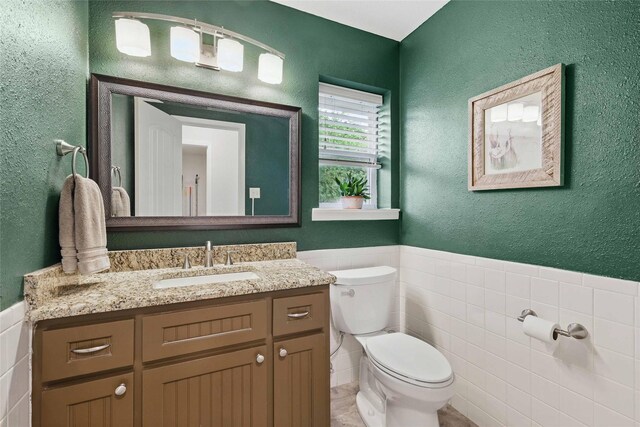 half bath featuring vanity, a textured wall, toilet, tile walls, and a wainscoted wall