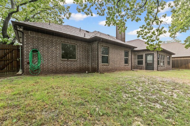 back of house featuring fence, roof with shingles, a chimney, brick siding, and a yard