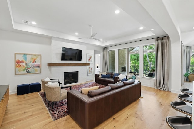 living area featuring ceiling fan, a fireplace, a tray ceiling, and light wood-style flooring