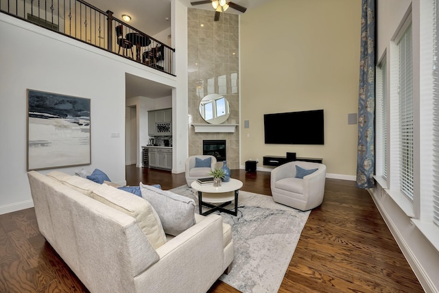 living room featuring ceiling fan, a towering ceiling, a tile fireplace, baseboards, and dark wood-type flooring