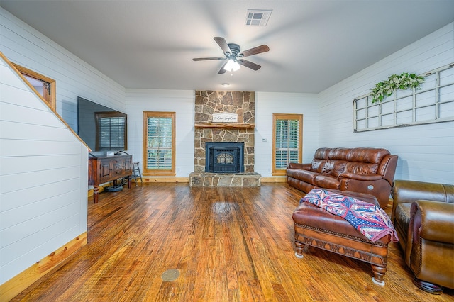 living area with wooden walls, wood-type flooring, a stone fireplace, a ceiling fan, and visible vents