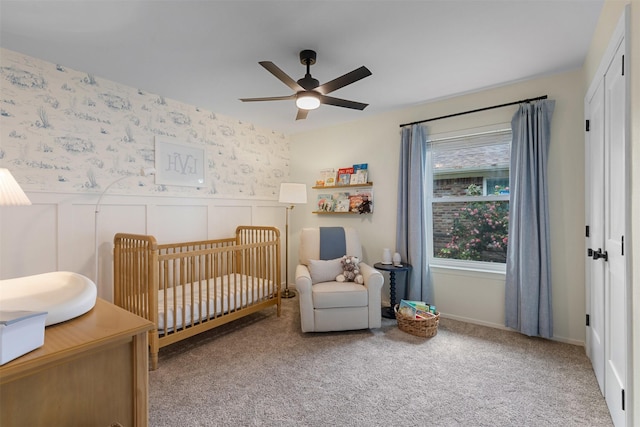 bedroom featuring a nursery area, ceiling fan, carpet, and a wainscoted wall