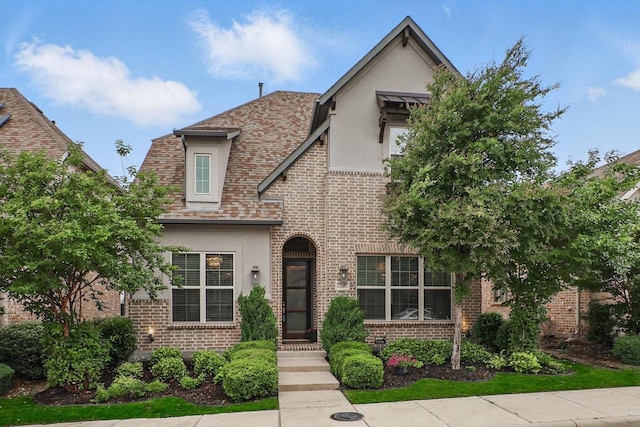 tudor home featuring brick siding, roof with shingles, and stucco siding