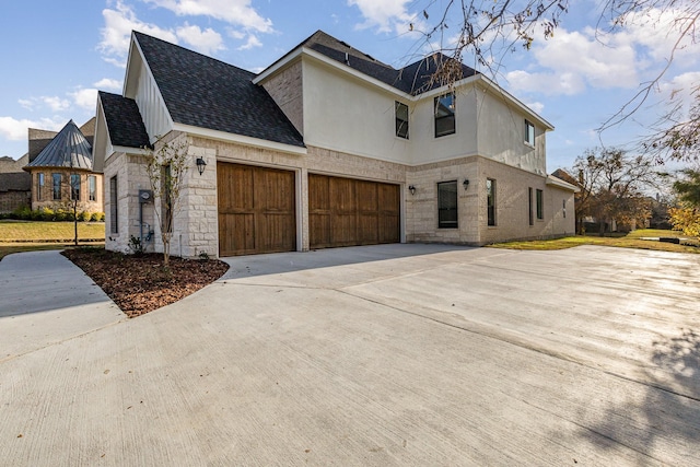 view of side of property featuring driveway, roof with shingles, a garage, stone siding, and stucco siding