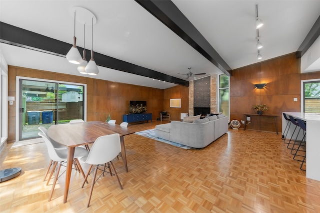 dining space featuring track lighting, wood walls, a brick fireplace, beamed ceiling, and ceiling fan