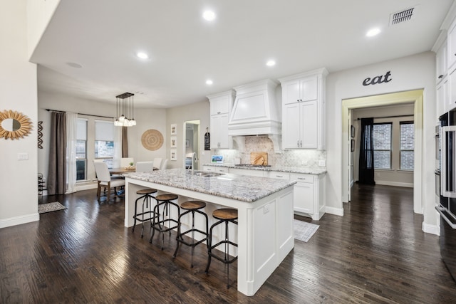 kitchen featuring an island with sink, white cabinets, custom range hood, and visible vents