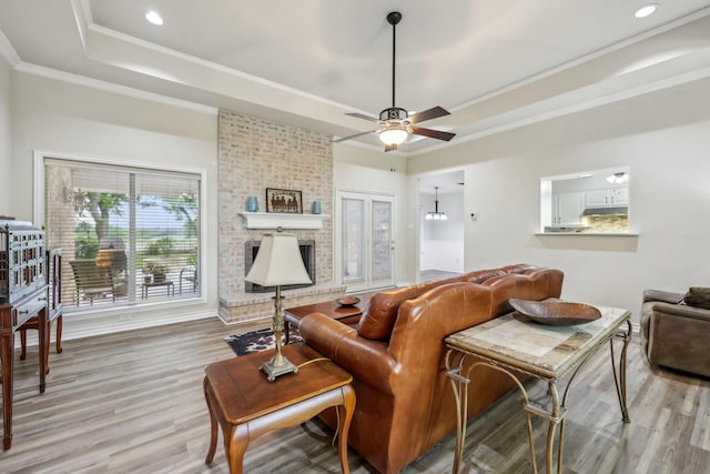 living room with ceiling fan, a fireplace, a raised ceiling, light wood-style floors, and ornamental molding