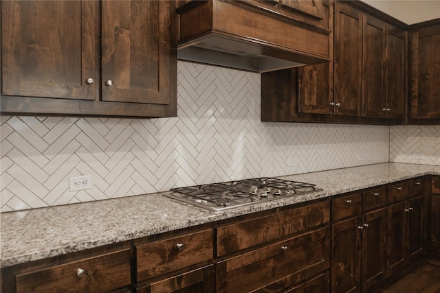 kitchen featuring decorative backsplash, stainless steel gas cooktop, light stone countertops, dark brown cabinets, and premium range hood
