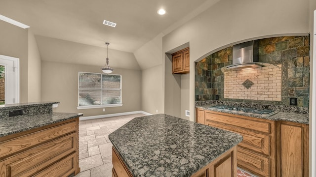 kitchen with brown cabinets, wall chimney range hood, stainless steel gas cooktop, dark stone countertops, and stone tile flooring