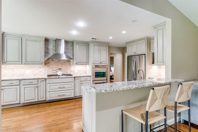 10907 Wonderland Trl, Dallas, TX 75229 - kitchen featuring a kitchen bar, wall chimney exhaust hood, a peninsula, stainless steel appliances, and light wood-style flooring