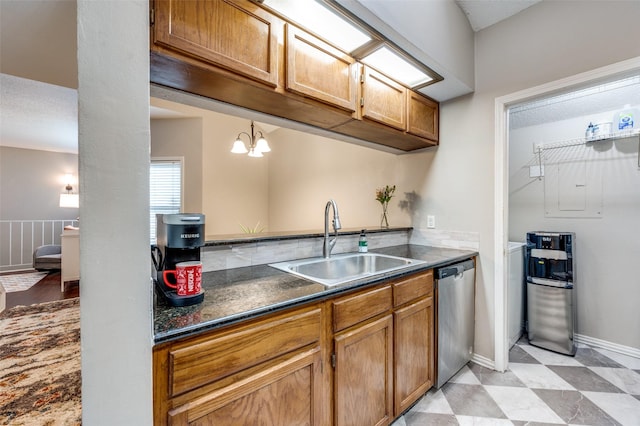 kitchen with stainless steel dishwasher, brown cabinetry, dark countertops, a sink, and a notable chandelier