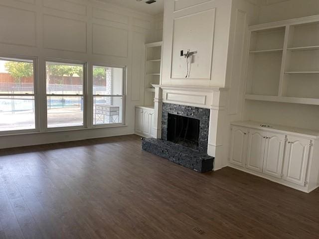 unfurnished living room featuring a stone fireplace, built in features, dark wood-type flooring, and a decorative wall
