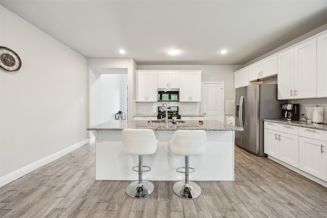kitchen featuring a kitchen island with sink, backsplash, appliances with stainless steel finishes, light wood-type flooring, and light stone counters
