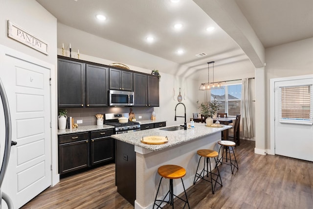 kitchen with a kitchen island with sink, a kitchen bar, appliances with stainless steel finishes, a sink, and dark wood-type flooring