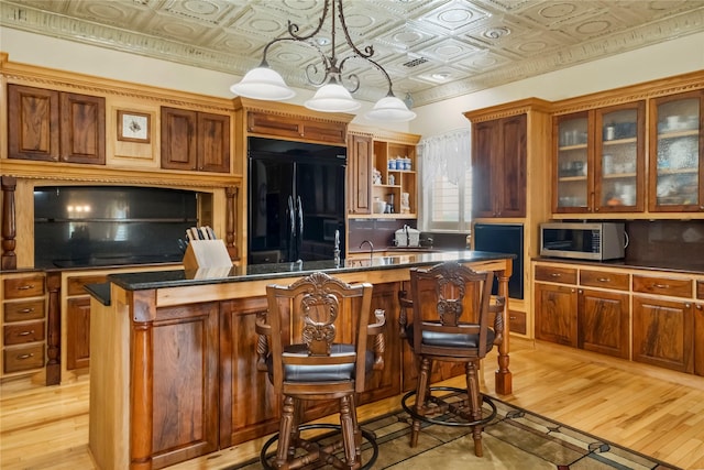 kitchen featuring an ornate ceiling, stainless steel microwave, black refrigerator, light wood-style flooring, and an island with sink